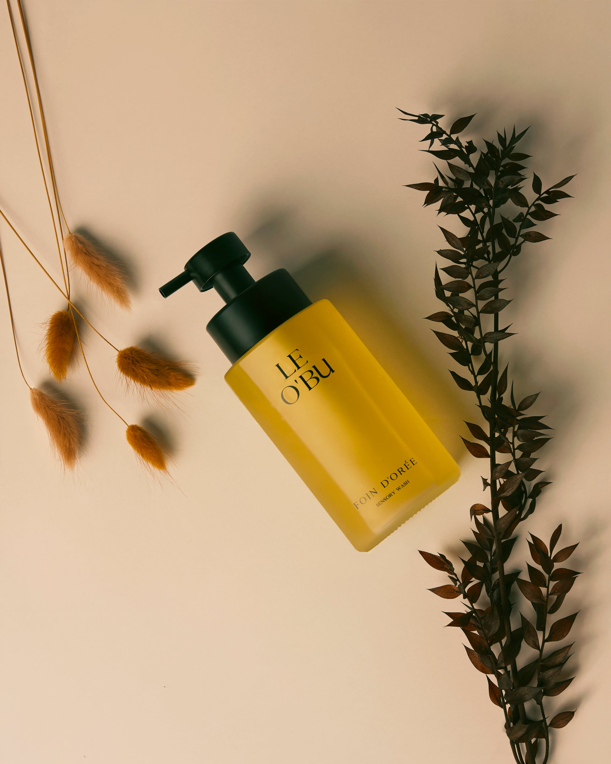Yellow bottle with black pump labeled 'LE OBU' on a beige background with plants.