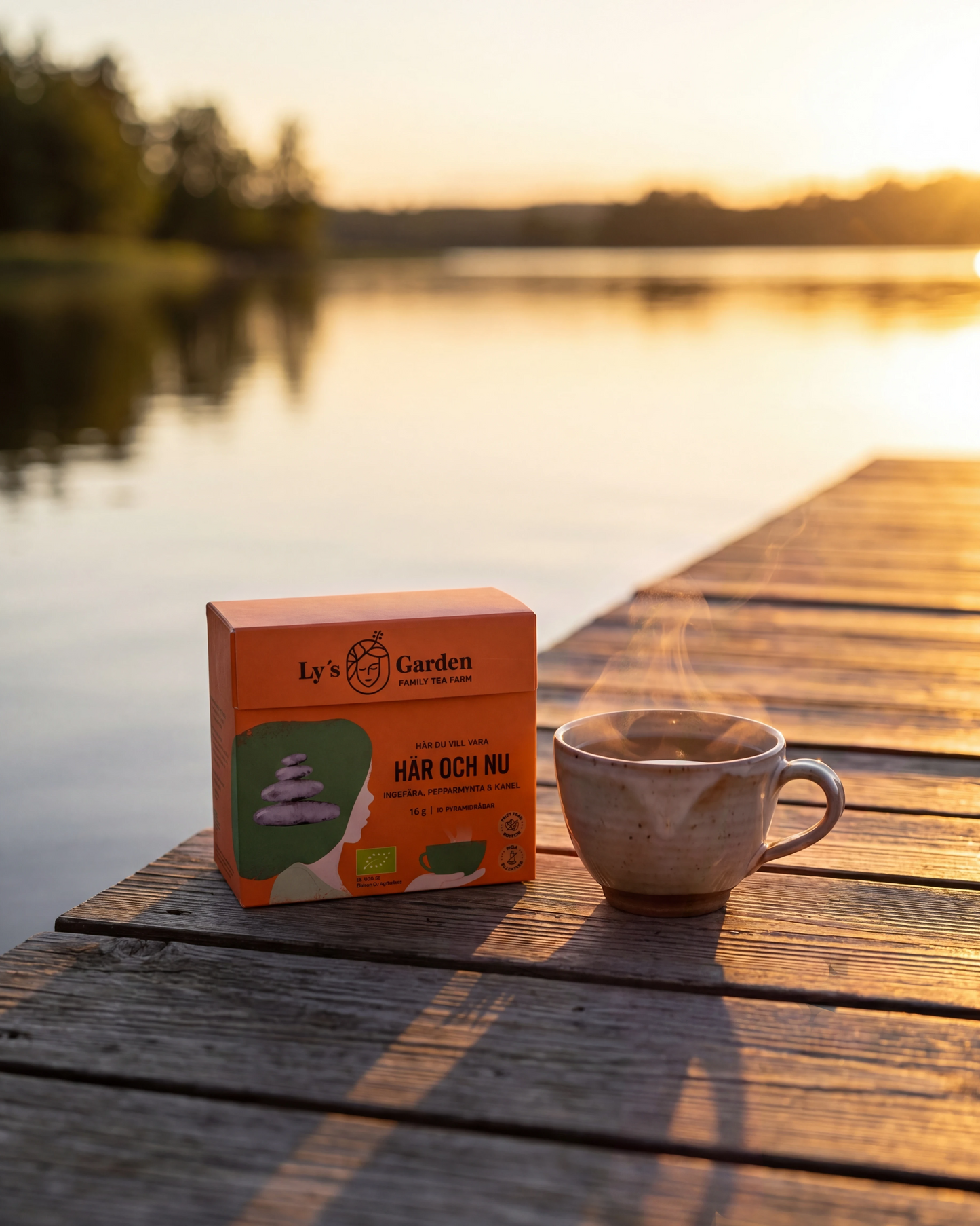 Box of Ly's Tea Garden tea and a steaming cup on a wooden dock by a lake at sunset.